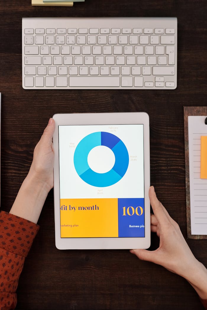A person holds a tablet showing a business graph next to a keyboard on a wooden desk.
